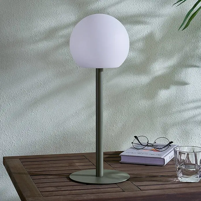 Lindby Tischleuchte Eleia - L x B x H: 16,5 x 16,5 x 44 cm, grün, weiß / opal - Olivgrüne Tischlampe mit kugelförmigem weißem Schirm, auf einem hölzernen Gartentisch mit Buch und Glas.