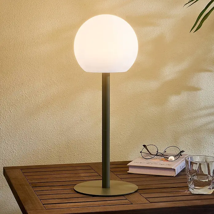 Lindby Tischleuchte Eleia - L x B x H: 16,5 x 16,5 x 44 cm, grün, weiß / opal - Moderne Tischlampe mit leuchtendem kugelförmigem weißen Schirm und dunklem grünem Stiel auf einem Holztisch neben einem Buch und Gläsern.