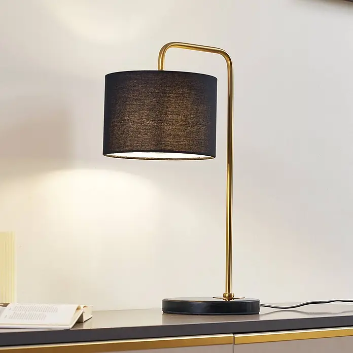 Lindby Tischleuchte Hillary - schwarz, schwarz, E27 - Eine stilvolle Tischlampe mit schwarzem Stoffschirm, goldenem Metallarm und dunklem Sockel, die ein warmes Licht abgibt.