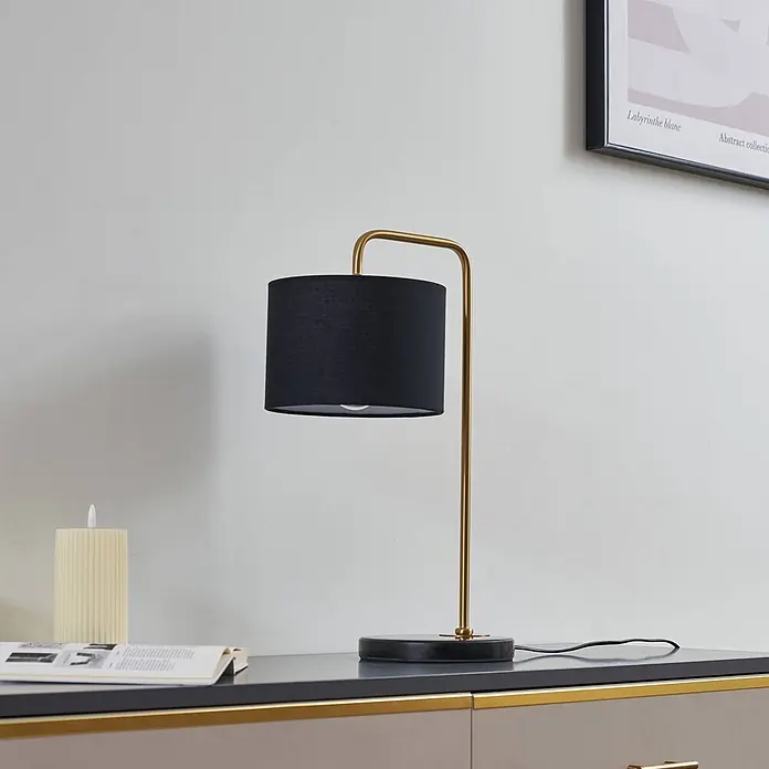 Lindby Tischleuchte Hillary - schwarz, schwarz, E27 - Eine Tischlampe mit schwarzem Schirm und goldfarbenem Ständer steht auf einer dunkelgrauen Fläche, neben einem Buch und einer Kerze.
