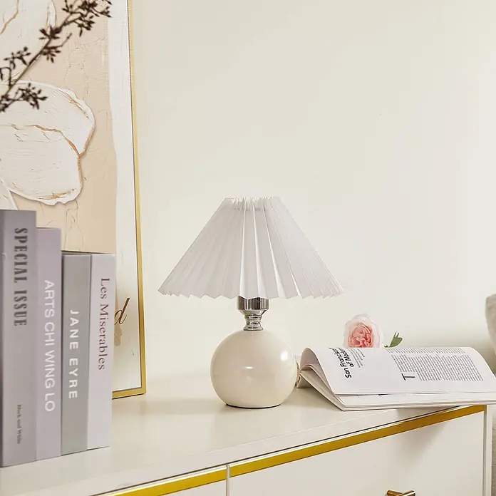 Lindby Tischleuchte Kerimi - L x B x H: 24 x 24 x 29 cm, weiß / opal, weiß / opal, E27 - Weiße Tischlampe mit plissiertem Schirm und rundem Keramikfuß, auf einer Kommode neben Büchern.