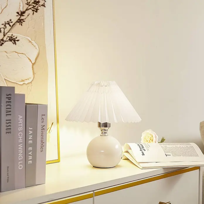 Lindby Tischleuchte Kerimi - L x B x H: 24 x 24 x 29 cm, weiß / opal, weiß / opal, E27 - Weiße Tischlampe mit Plisseeschirm und rundem Keramikfuß auf einer Kommode neben Büchern.