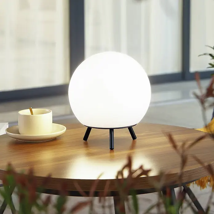 Lindby Tischleuchte Aurilia - schwarz, weiß / opal - Eine weiße kugelförmige Tischleuchte mit schwarzem Stativfuß steht auf einem Holztisch neben einer Tasse und schafft eine gemütliche Atmosphäre.
