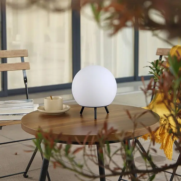 Lindby Tischleuchte Aurilia - schwarz, weiß / opal - Eine weiße, kugelförmige LED-Lampe mit schwarzen Stativbeinen steht auf einem runden Holztisch auf einem Balkon.