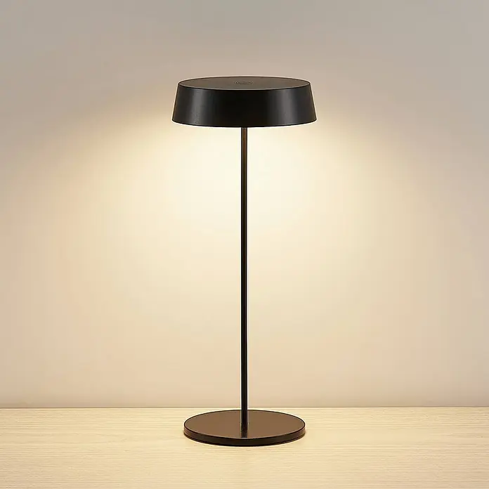 Lucande Tischleuchte Tibia - L x B x H: 12 x 12 x 29,5 cm, schwarz, schwarz - Moderne schwarze Tischleuchte mit rundem Fuß und Schirm, die warmes Licht auf eine Holzfläche wirft.