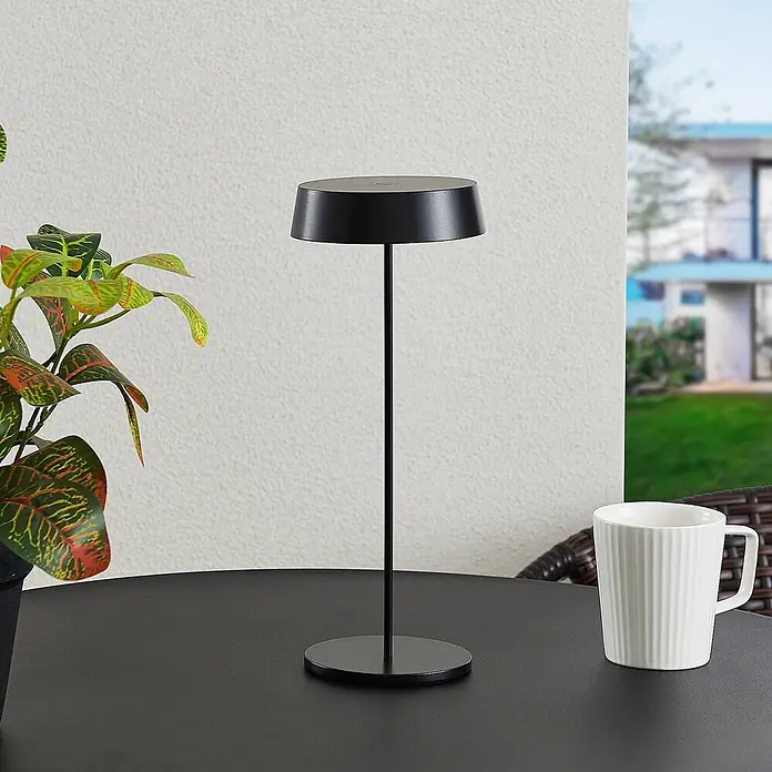 Lucande Tischleuchte Tibia - L x B x H: 12 x 12 x 29,5 cm, schwarz, schwarz - Eine minimalistische schwarze tragbare Tischleuchte auf einem Gartentisch, neben einer Topfpflanze und einer weißen Tasse.