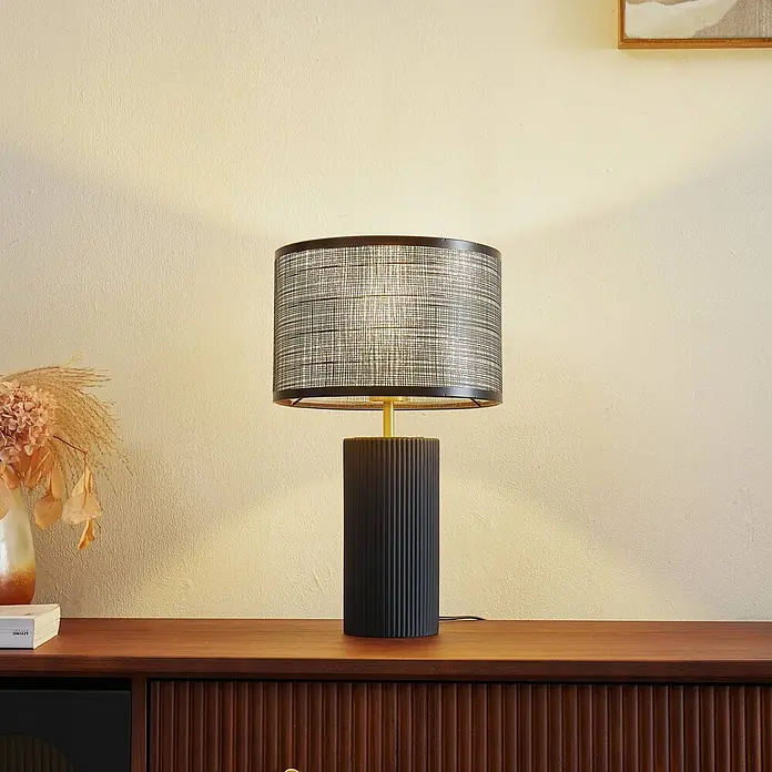 Lindby Tischleuchte Sorscha - schwarz, schwarz, E27 - Schwarze geriffelte Tischlampe mit Goldstiel und strukturiertem schwarzem Lampenschirm, beleuchtet auf einem Holzschrank.