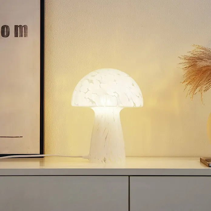 Lindby Tischleuchte Hubi - L x B x H: 22 x 22 x 28 cm, weiß / opal, weiß / opal, E27 - Eine beleuchtete Pilz-Tischlampe aus weißem, gesprenkeltem Glas steht auf einer weißen Kommode.