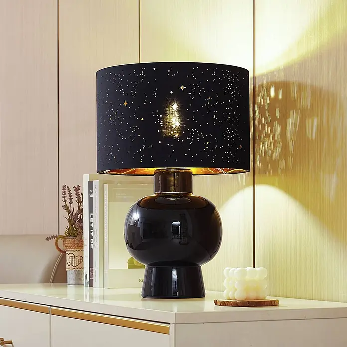 Lindby Tischleuchte Arved - L x B x H: 30 x 30 x 46 cm, schwarz, schwarz, E27 - Eine schwarze Tischlampe mit glänzendem Keramikfuß und schwarzem Lampenschirm mit goldenen Sternenausschnitten, der Sternenmuster an die Wand wirft.