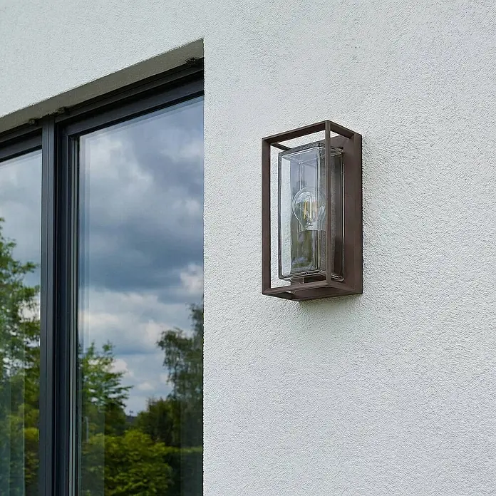 Arcchio Außenwandleuchte Ismera - braun / rost, braun / rost, IP65 - Braune Outdoor-Wandleuchte mit Klarglasschirm und sichtbarer Glühbirne, an einer strukturierten Wand neben einem Fenster montiert.