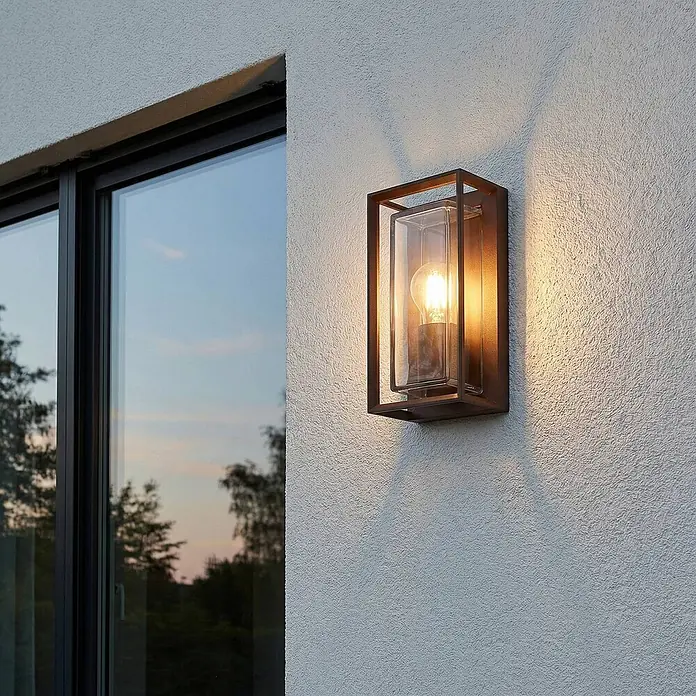 Arcchio Außenwandleuchte Ismera - braun / rost, braun / rost, IP65 - Rechteckige Außenwandleuchte mit dunklem Metallrahmen, Klarglas und sichtbarer Glühbirne, beleuchtet an einer strukturierten Wand neben einem Fenster.