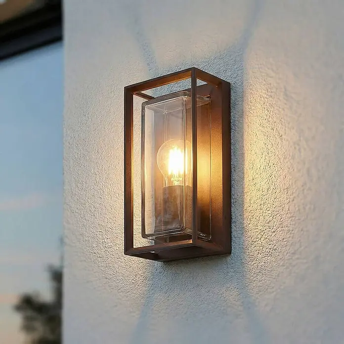Arcchio Außenwandleuchte Ismera - braun / rost, braun / rost, IP65 - Dunkelbraune Metall-Außenwandleuchte mit klarem Glasschirm, an strukturierter Außenwand montiert und beleuchtet.