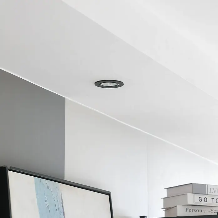 Arcchio Deckenstrahler Zarik - schwarz, schwarz - Ein schwarzer Einbau-Deckenstrahler beleuchtet einen modernen Raum mit weißen und grauen Wänden, über Büchern und einem Gemälde.