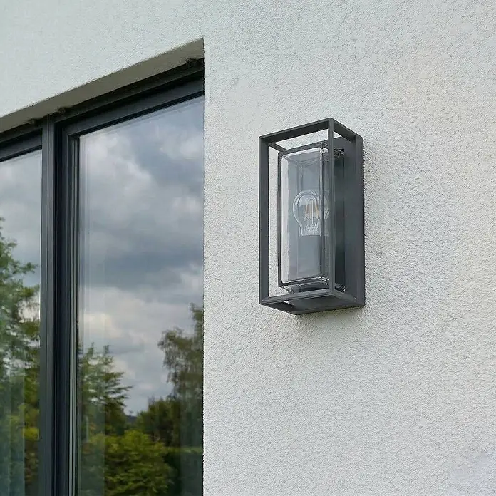 Arcchio Außenwandleuchte Ismera - schwarz, schwarz, IP65 - Dunkelgraue rechteckige Außenwandleuchte mit Klarglas und sichtbarer Filamentbirne, an weißer Strukturwand neben Fenster montiert.