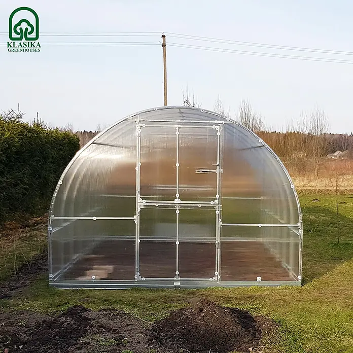 Klasika Greenhouses Gewächshaus Tube Klasika - Außenmaß inkl. Dachüberstand (B x T): 300 x 1.000 cm, Material Seitenwand: Kunststoff, 4 mm, Silbergrau - Gewölbtes, transparentes Gewächshaus mit Metallrahmen, draußen auf Rasen mit etwas Erde aufgestellt.