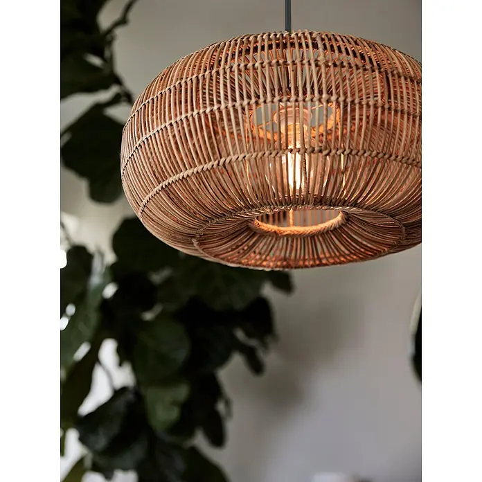 PR Home Pendelleuchte Rattan Outdoor - 15 W, E27 - Runde Hängeleuchte aus Rattan, beleuchtet, hängt in einem Raum mit unscharfer grüner Pflanze.
