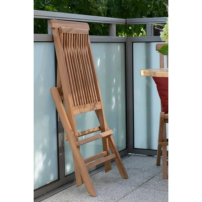 Bene Living Gartentisch-Set Langton & Tisch Sutton - Gesamtumfang: 3 -tlg., Teakholz, Klappbar - Ein zusammengeklappter Holzstuhl in Naturholzoptik lehnt an einem Balkongeländer.