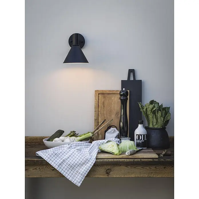 PR Home Außenwandleuchte Mini-Tripp - 15 W, IP44 - Eine schwarze Wandlampe beleuchtet eine rustikale Küchentheke mit frischem Gemüse und Kochzubehör.