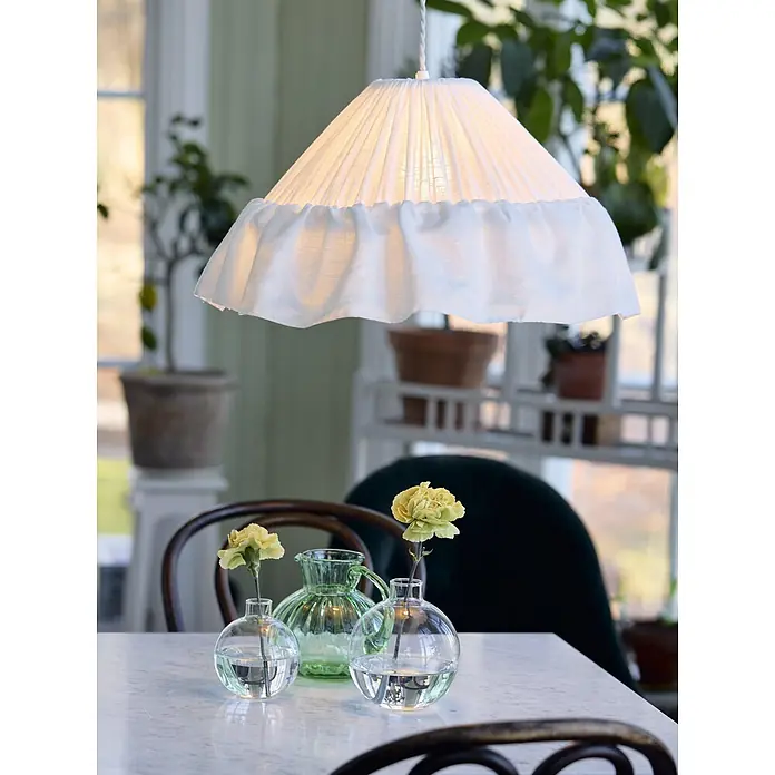 PR Home Pendelleuchte Stina - 15 W, E27 - Weiße plissierte Stoff-Pendelleuchte mit Rüschenkante, hängt über einem Tisch in einem hellen Raum.