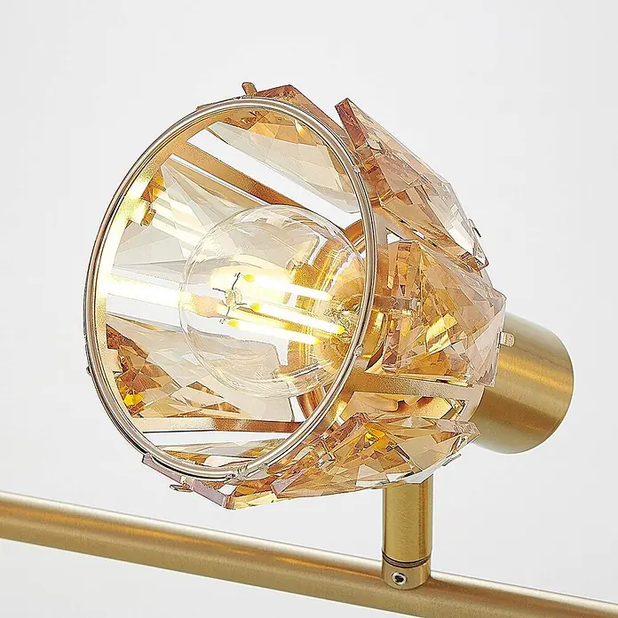 Lindby Deckenleuchte Kosta - L x B x H: 70 x 15,5 x 18 cm, messing / gold, klar / transparent, E14 - Nahaufnahme eines goldenen Strahlers mit facettierten bernsteinfarbenen Kristallelementen und einer beleuchteten LED-Glühbirne.