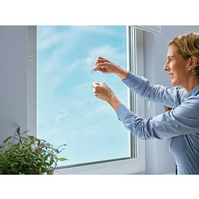 Tesa Colgador adhesivo - Transparente, Carga soportada: 0,2 kg, 5 ud. - Mujer aplicando una lámina para ventanas para aumentar la privacidad y reducir el calor en la habitación.
