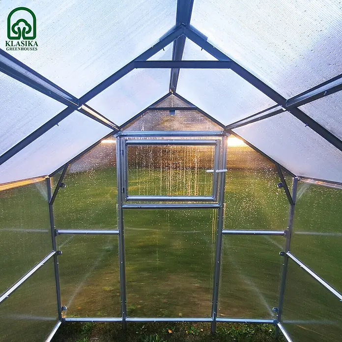 Klasika Greenhouses Gewächshaus Bernard Klasika - Außenmaß inkl. Dachüberstand (B x T): 235 x 800 cm, Material Seitenwand: Kunststoff, 6 mm, Silbergrau - Inneransicht eines Klasika Gewächshauses mit transparenten Platten und Metallrahmen, offenem Lüftungsfenster und Sonnenlicht. Wassertropfen sichtbar.