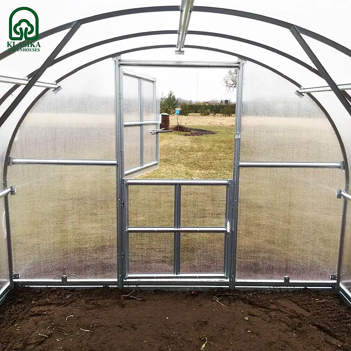 Klasika Greenhouses Gewächshaus Arch Klasika - Außenmaß inkl. Dachüberstand (B x T): 300 x 1.200 cm, Material Seitenwand: Kunststoff, 6 mm, Silbergrau - Innenansicht eines bogenförmigen Gewächshauses mit transparenten Wänden und silbernem Metallrahmen, Tür zu einem Feld geöffnet.