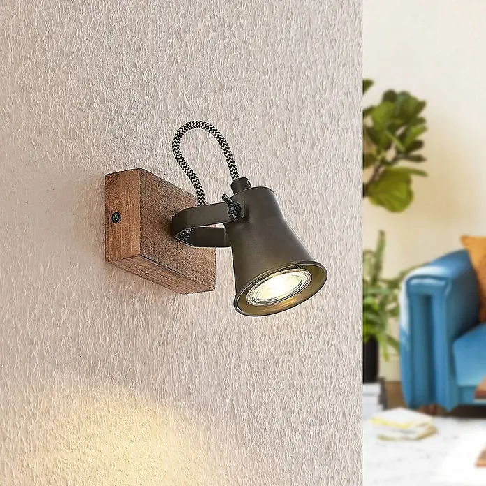 Lindby Deckenstrahler Grandesa - Holz dunkel, schwarz, GU10 - Rustikaler Wandstrahler mit dunklem Metallkopf und geflochtenem Kabel, auf Holzblock montiert, wirft warmes Licht an eine strukturierte Wand.