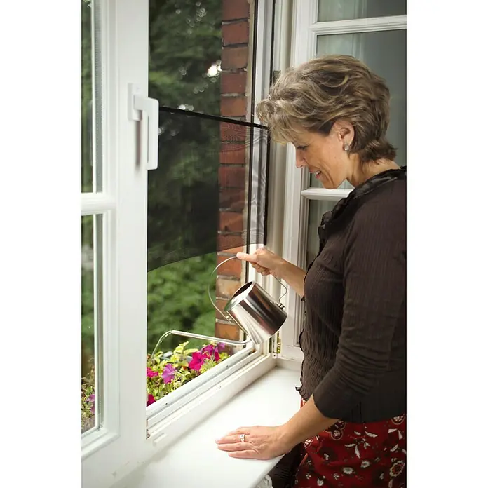 Tesa Insect Stop Mosquitera para ventana - An x Al: 150 x 130 cm, Montaje de la mosquitera: Enganche - Una mujer riega flores en una jardinera con una jarra brillante y metálica que refleja la luz.