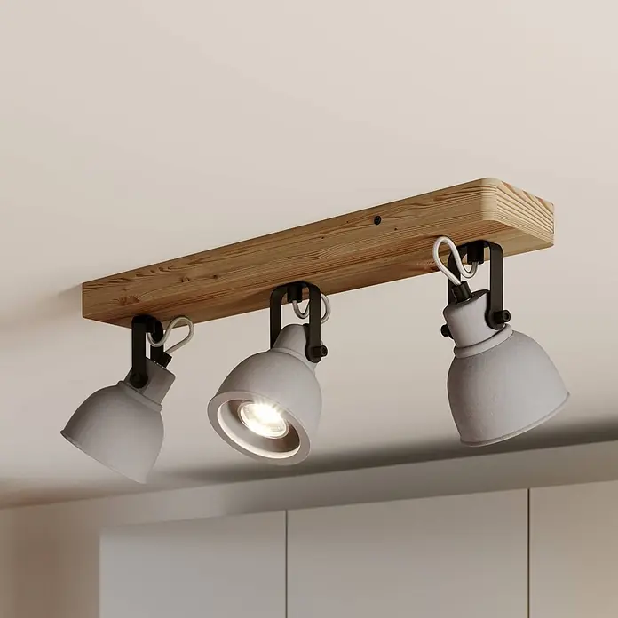 Lindby Deckenstrahler Mitis - Holz hell, alu / grau / zink, GU10 - Deckenleuchte mit drei grauen Beton-Spots auf einem Holzbalken. Ein Spot ist an und beleuchtet den Raum.