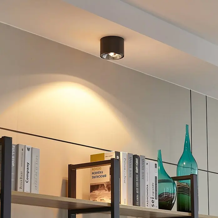 Arcchio Deckenstrahler Mabel - alu / grau / zink, alu / grau / zink, GU10 - Dunkler zylindrischer Deckenstrahler wirft warmes Licht auf eine Wand über einem Bücherregal mit Büchern und Vasen.