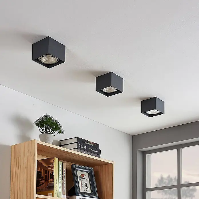 Arcchio Deckenstrahler Mabel - alu / grau / zink, alu / grau / zink, GU10 - Drei schwarze, würfelförmige Deckenstrahler an einer weißen Decke über einem Holzregal.