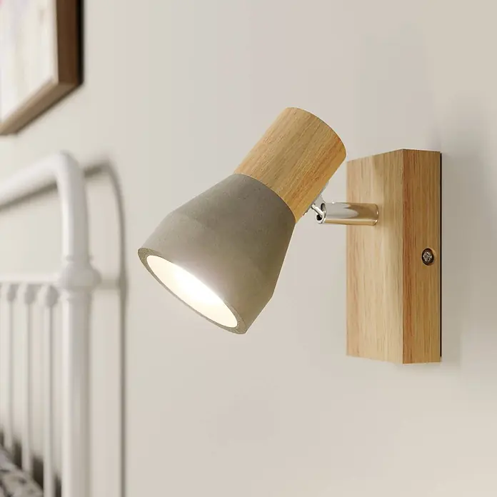 Lindby Deckenstrahler Filiz - Holz hell, alu / grau / zink, E14 - Verstellbare Wandleuchte mit grauem Betonschirm und hellem Holzsockel, neben einem weißen Bett platziert.