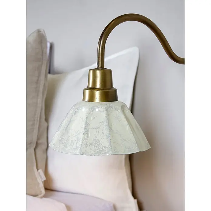 PR Home Pendelleuchte Ester - 25 W, E27 - Messing-Wandleuchte mit cremefarbenem, gemustertem Glasschirm, über Kissen am Bett positioniert.