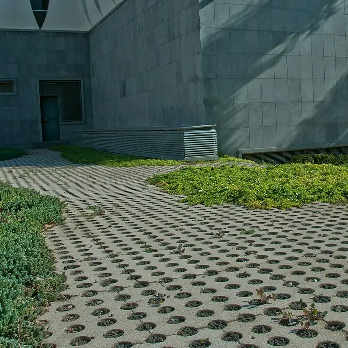 Adoquín para jardín - 60 x 36 x 8 cm, Gris, Hormigón - Adoquines de hormigón con agujeros que permiten el crecimiento del césped, colocados frente a un edificio. Las piedras forman un camino.