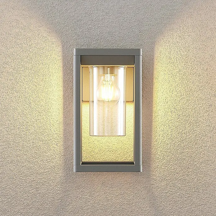 Lindby Wandleuchte aussen in silber, Aussenlampe Wand -  - Graue rechteckige Außenwandleuchte mit klarem Glaszylinder und warmem Filamentlicht, das nach oben und unten auf eine strukturierte Wand strahlt.