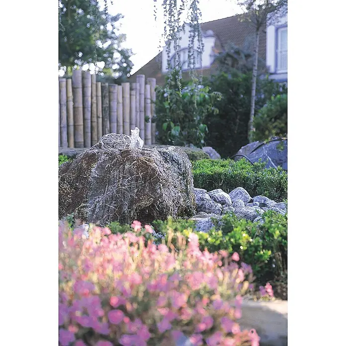 Oase Statuen-Zimmerbrunnenpumpe Aquarius Universal 2000 - 25 W, Max. Förderhöhe: 200 cm - Gartenbrunnen aus Fels mit sprudelndem Wasser, umgeben von grünen Pflanzen und verschwommenen rosa Blüten.