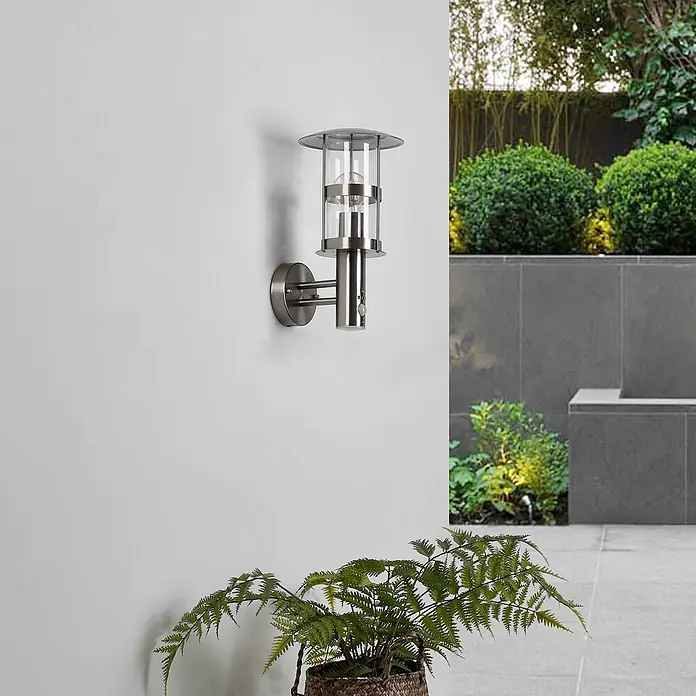 Lindby Außenwandleuchte Noemi - alu / grau / zink, alu / grau / zink, IP44 - Gebürstete Edelstahl-Außenwandleuchte im Laternenstil mit klarem Glas und Bewegungssensor an einer hellen Außenwand.