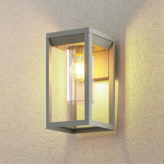 Lindby Wandleuchte aussen in silber, Aussenlampe Wand -  - Anthrazitfarbene rechteckige Wandleuchte mit Klarglaszylinder und beleuchteter Glühbirne an strukturierter Wand.
