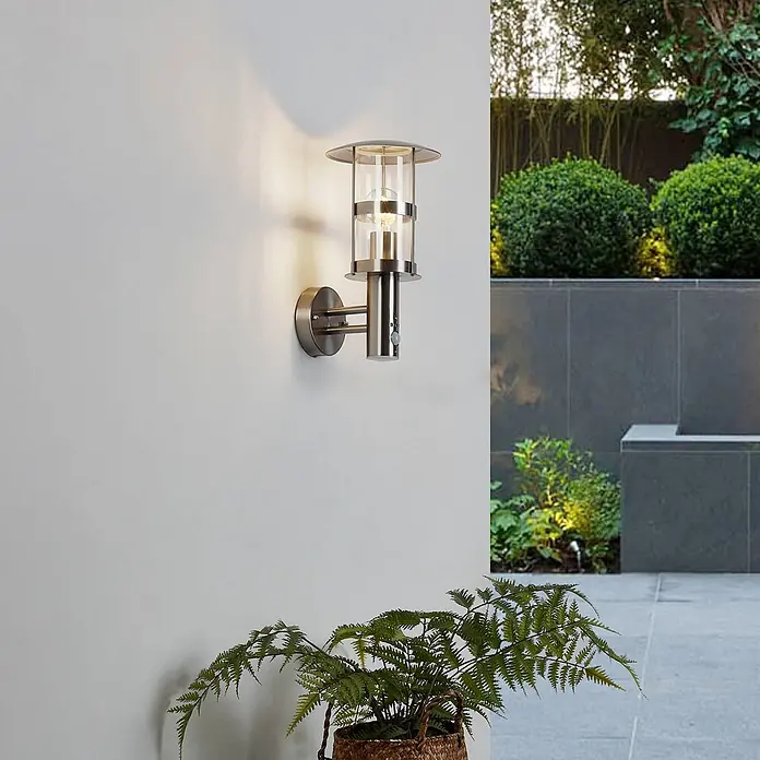 Lindby Außenwandleuchte Noemi - alu / grau / zink, alu / grau / zink, IP44 - Gebürstete Metall-Außenwandleuchte mit Klarglas und integriertem Bewegungssensor, beleuchtet eine Terrassenwand.