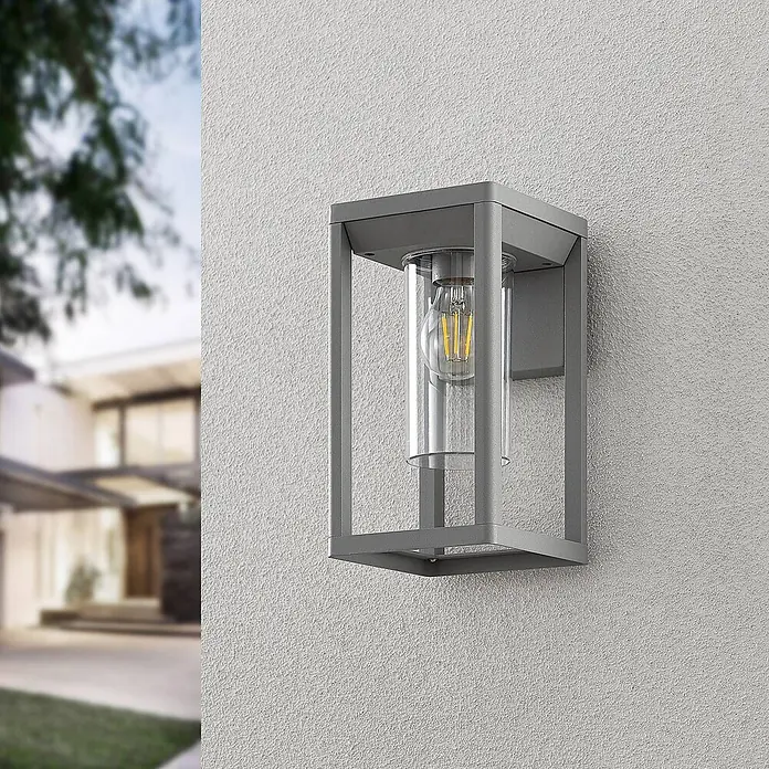 Lindby Wandleuchte aussen in silber, Aussenlampe Wand -  - Moderne graue Außenwandleuchte mit klarem Glaszylinder und LED-Filamentlampe, an einer texturierten Hauswand montiert.