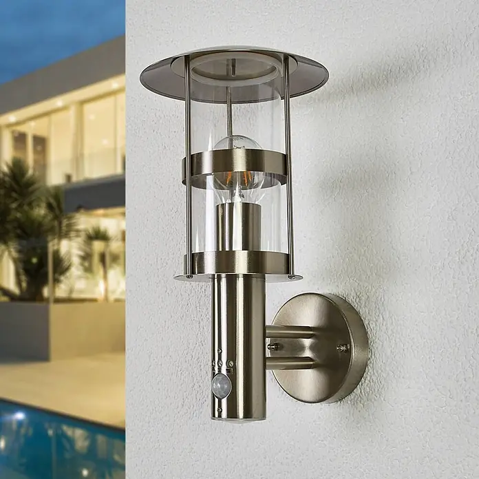 Lindby Außenwandleuchte Noemi - alu / grau / zink, alu / grau / zink, IP44 - Edelstahl-Außenwandleuchte mit klarem Glas und Bewegungsmelder, an einer Hauswand vor einem modernen Gebäude.
