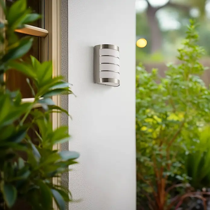 Lindby Außenwandleuchte Noyan - alu / grau / zink, alu / grau / zink, IP44 - Gebürstete silberne Außenwandleuchte mit weißen Diffusorstreifen an einer Außenwand neben einem Fenster, umgeben von grünen Pflanzen.
