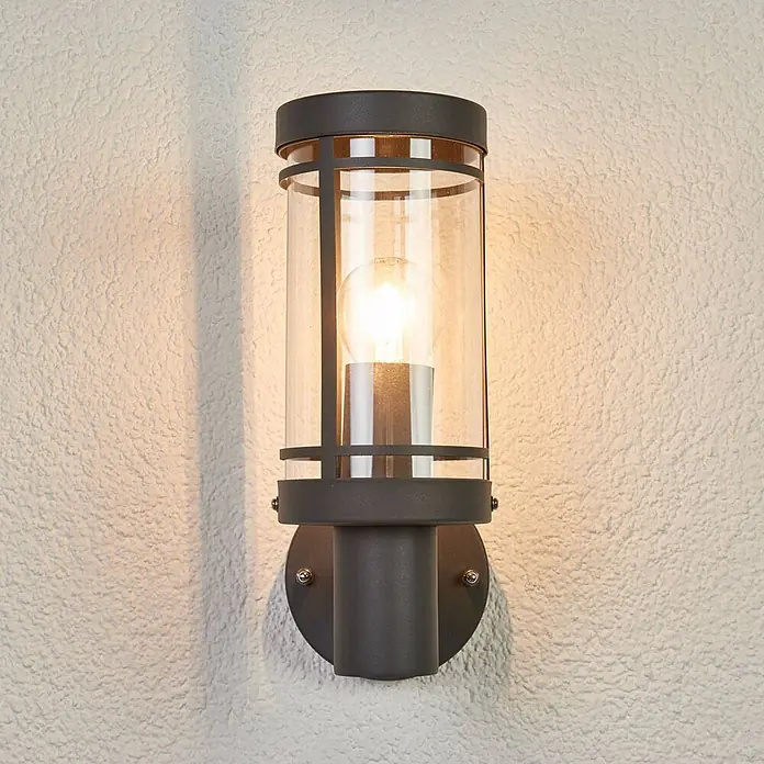 Lindby Außenwandleuchte Djori - alu / grau / zink, IP44 - Dunkelgraue zylindrische Außenwandleuchte mit Klarglas und sichtbarer Glühbirne, an einer strukturierten Wand montiert.
