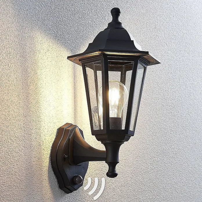 Lindby Außenwandleuchte Nane - schwarz, schwarz, IP44 - Schwarze Außenwandleuchte aus Metall mit Klarglas, sichtbarer Glühbirne und integriertem Bewegungssensor, die eine strukturierte Wand beleuchtet.