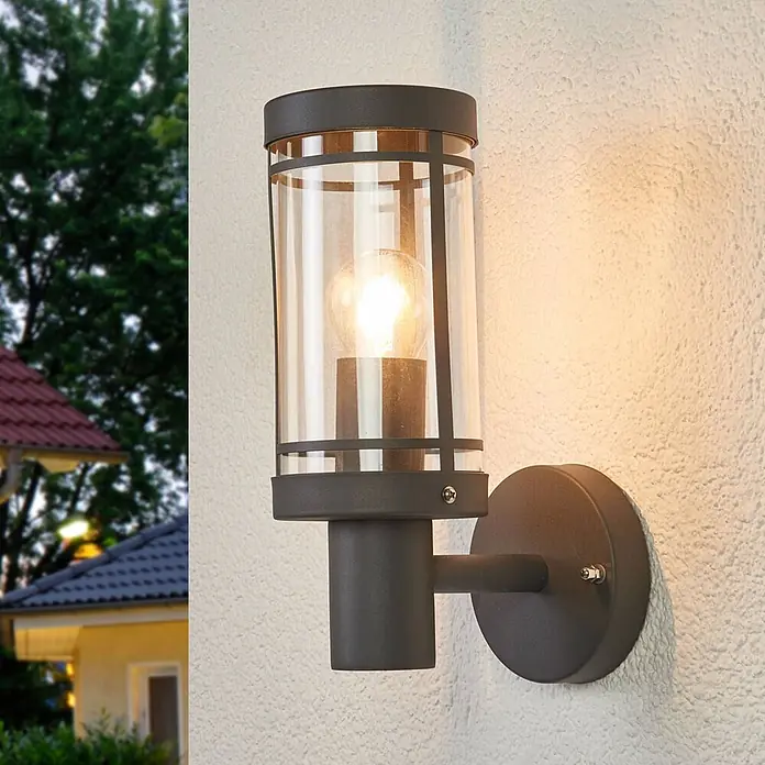 Lindby Außenwandleuchte Djori - alu / grau / zink, IP44 - Dunkelgraue Außenwandleuchte mit zylindrischem Klarglasschirm, an einer strukturierten Wand montiert.