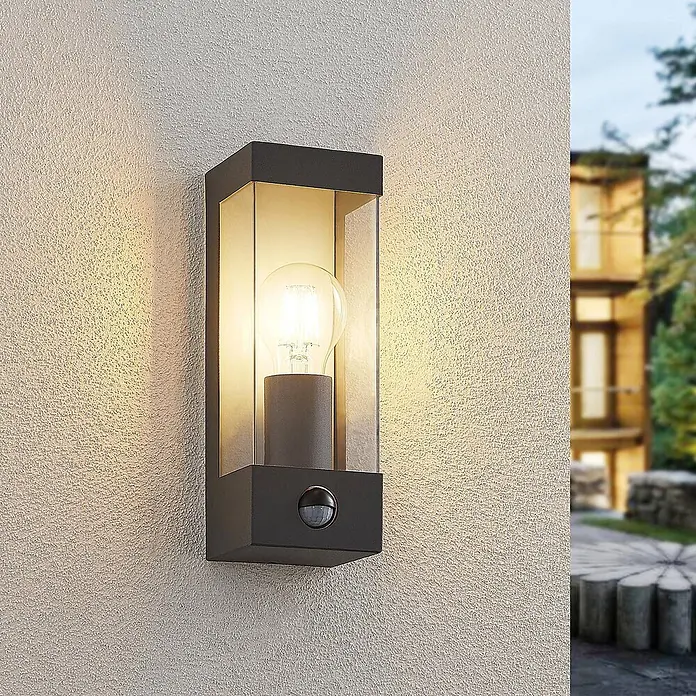Lindby Außenwandleuchte Tilian - alu / grau / zink, alu / grau / zink, IP44 - Dunkelgraue Außenwandleuchte aus Metall und Glas mit sichtbarer Glühbirne und Bewegungssensor an einer strukturierten Wand.