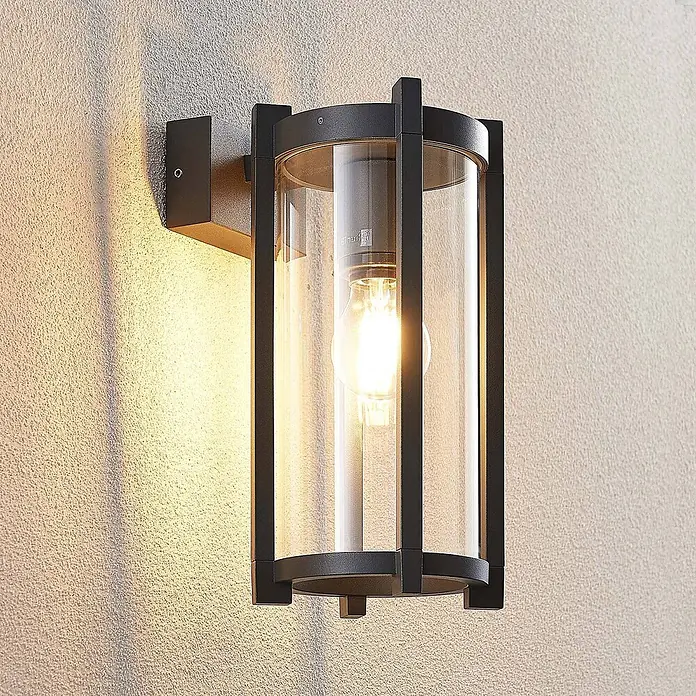 Lucande Außenwandleuchte Brienne - schwarz, IP54 - Schwarze Außenwandleuchte aus Metall mit klarem Glaszylinder und sichtbarer Glühbirne, an strukturierter Wand montiert.