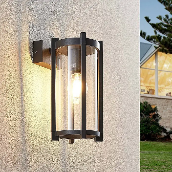 Lucande Außenwandleuchte Brienne - schwarz, IP54 - Schwarze Außenwandleuchte mit klarem Glaszylinder und beleuchteter Glühbirne, an einer strukturierten Wand montiert.