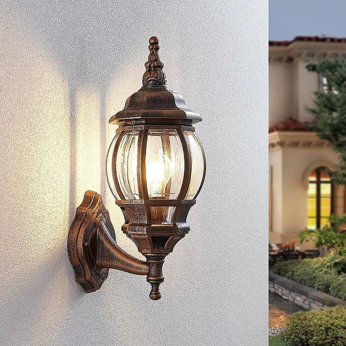 Lindby Außenwandleuchte Nadesha - messing / gold, messing / gold, IP44 - Verzierte Wandlaterne aus gealterter Bronze mit Klarglas, leuchtet eine helle Wand neben einem Haus und Garten.
